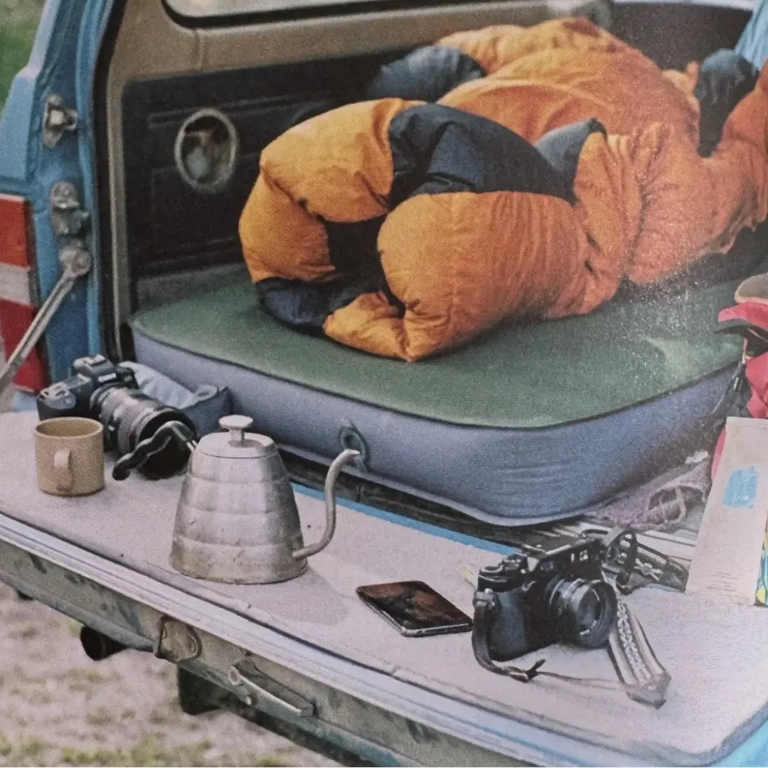 Vintage outdoor camping gear displayed in the back of a classic blue vehicle, featuring an orange sleeping bag, camping kettle, coffee mug, and retro film cameras — part of the Foraged Finds vintage outdoor collection.”