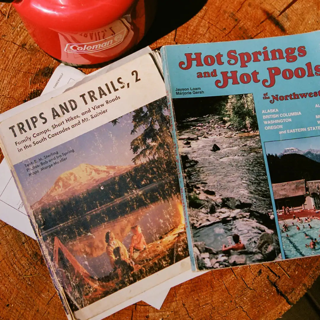 Two well-worn vintage outdoor guidebooks rest on a rustic wooden surface beside a red Coleman mug. The left book, titled “Trips and Trails, 2,” features a cover photo of campers by a lake with Mount Rainier in the background at sunset. The right book, “Hot Springs and Hot Pools of the Northwest,” displays images of rivers, hot springs, and a pool surrounded by mountains. A few sheets of paper peek out from underneath the books, adding to the casual, adventure-ready scene.