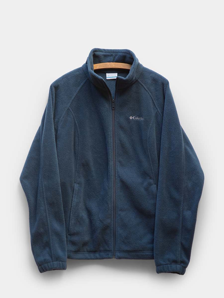 front-facing product photo of a deep blue columbia full zip fleece jacket on a wooden hanger against a light gray background. soft fleece texture with stand collar, zip closure, and side pockets visible. clean, minimal presentation with no visible wear.