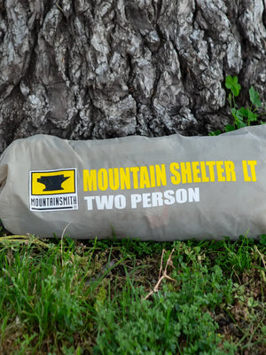 Mountainsmith Mountain Shelter LT photographed in carrying bag lying on green grass and tree stump in background