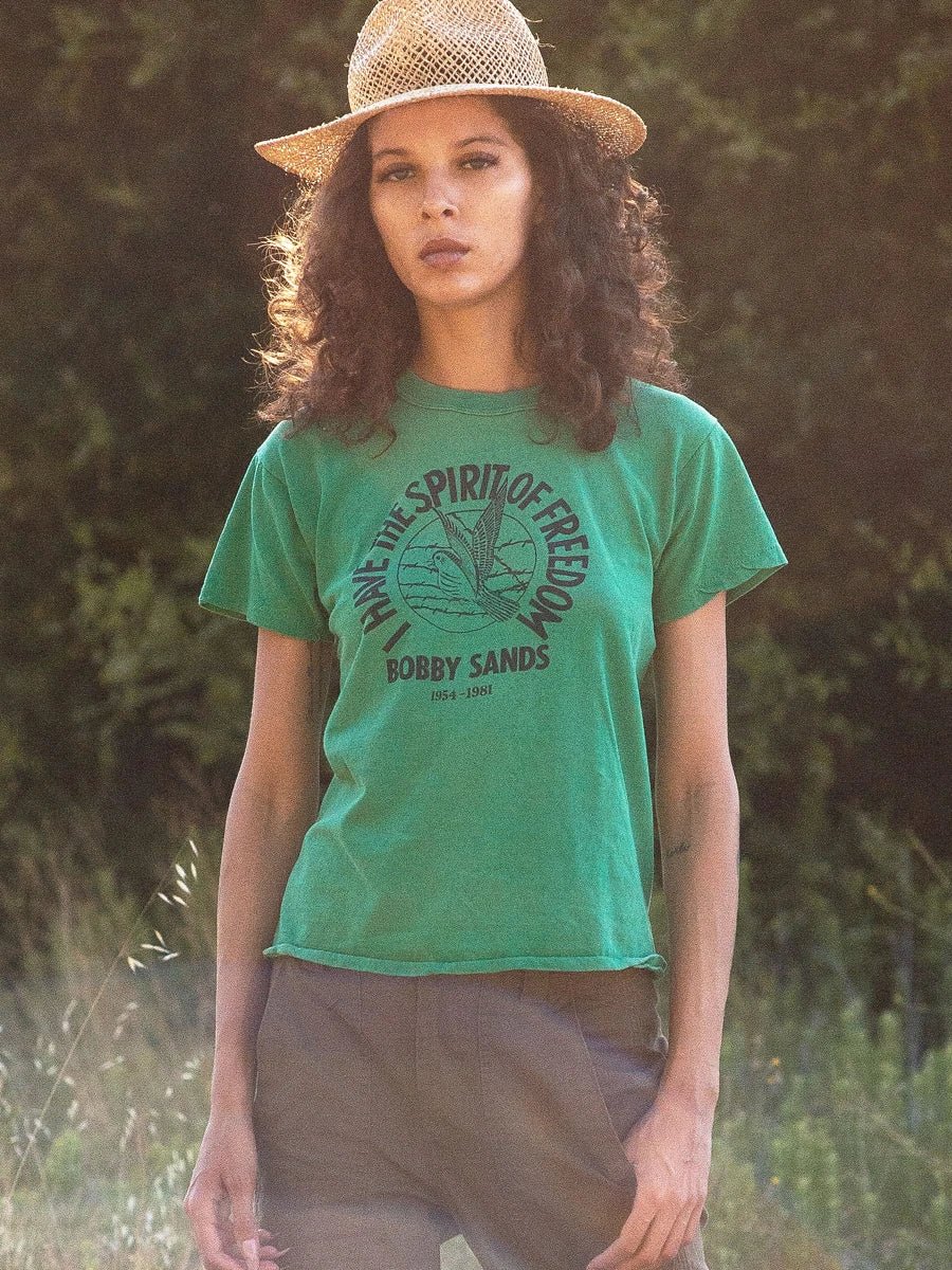 Model is in a nature setting wearing a green vintage tee, modern sun hat and brown pants in a relaxed pose 
